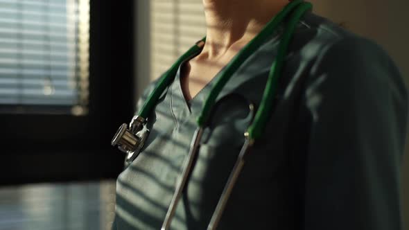 Closeup of Unrecognizable Female Doctor in Blue Green Uniform Removes Stethoscope From Neck Standing