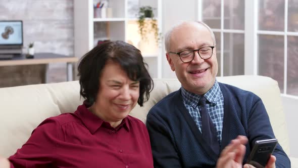 Happy Elderly Couple Sitting on Sofa Holding Smartphone