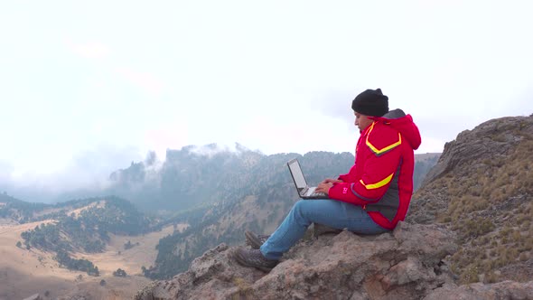 digital nomad, young latin man working on his laptop sitting on a rock in the mountain, backpacking