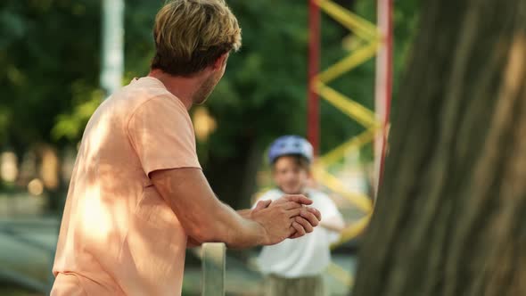 A pleased father is standing in the stands and watching his son riding a penny board at the park at