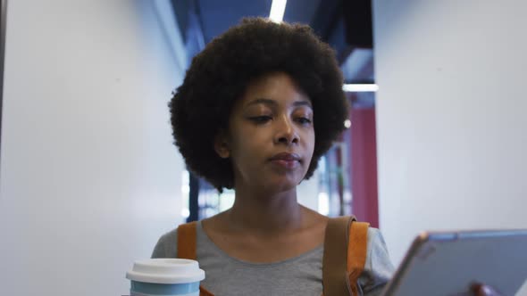 Mixed race businesswoman walking using digital tablet and drinking a cup of coffee in modern office