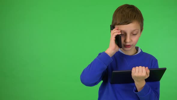 A Young Cute Boy Holds a Tablet and Talks on a Smartphone - Green Screen Studio