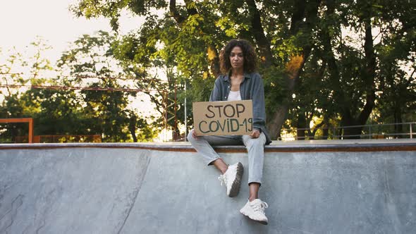 Afroamerican Girl in Casual Clothes is Showing Cardboard Poster with Inscription Stop Covid19