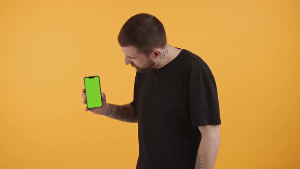 Attractive Bearded Man Holds Smartphone with Green Display Shows Thumbs Up Looking at the Camera