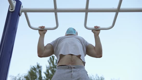 The athlete performs exercises on the horizontal bar in a medical mask.