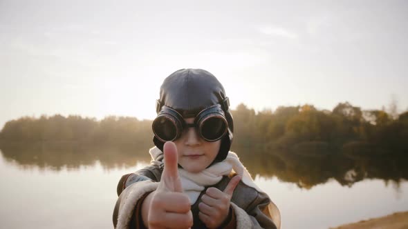 Little Thoughtful Aviator Boy Looking at Camera, Wearing Pilot Glasses and Showing Thumbs Up at