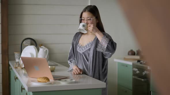 Concentrated Young Asian Woman Working Online in the Morning in Kitchen Drinking Coffee