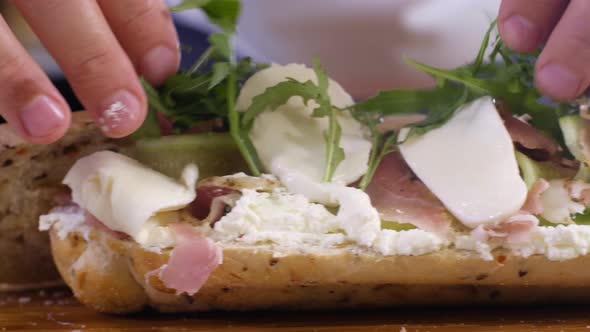 Young chef puts thin green fresh arugula lettuce leaves on a sandwich in home