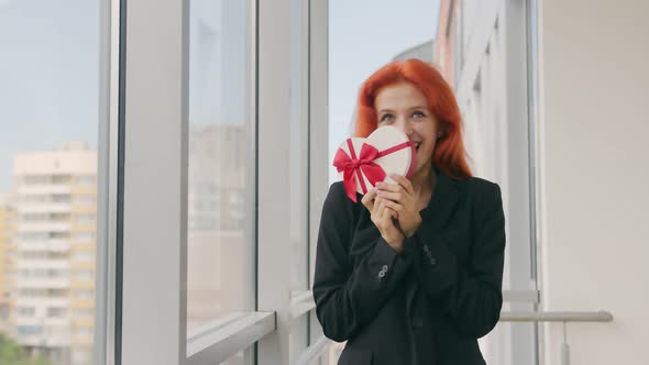 The Woman Received a Gift Box in the Form of a Heart. Happy Woman with Red Hair Rejoices at the Gift