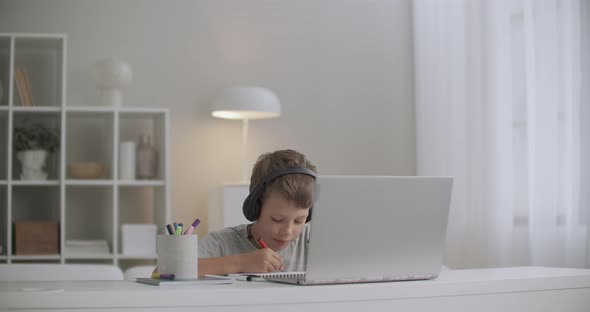 School Boy Is Listening Fairytale Through Headphones By Notebook and Drawing, Sitting at Table Alone