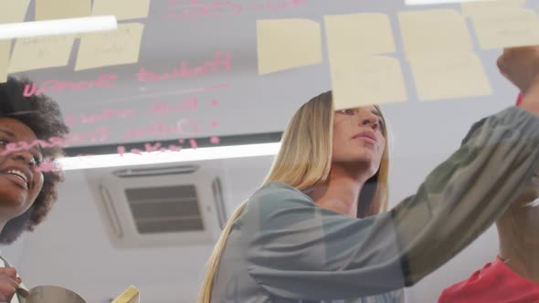 Three diverse businesswomen brainstorming writing memo notes on glass wall in office