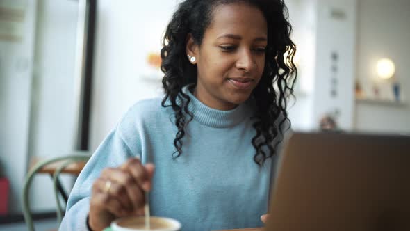 Pretty African woman wearing blue sweater working by laptop and drinking coffee