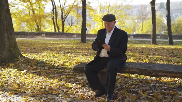 Happy Senior Man Browses the Tablet Screen When Resting on Bench in Autumn Park