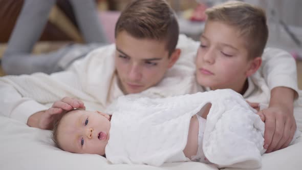 Relaxed Adorable Baby Girl Lying on White Bed with Blurred Boys Stroking Head Admiring Newborn Child