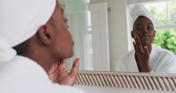 African american woman in bathrobe applying face cream while looking in the mirror at home