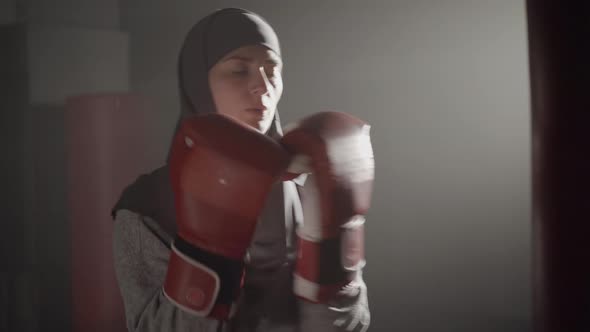 Concentrated Muslim Sportswoman in Hijab Fighting Punching Bag in Foggy Backlight. Portrait of