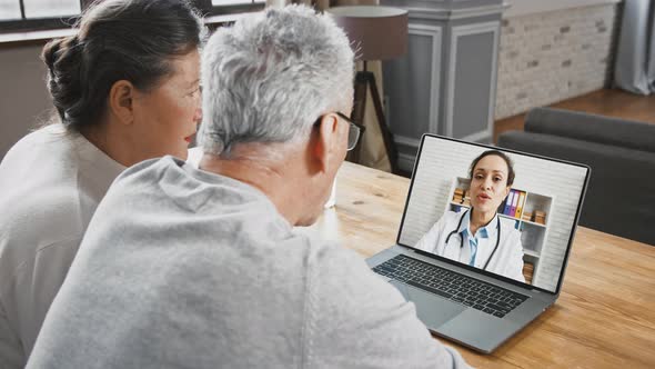 Back View of Aged Couple Looking at Screen of Laptop and Talking to Doctor Woman Using Online Video