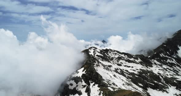 Backward Aerial Top View Over Cloudy Rocky Snowy Mountain in Sunny Day with Clouds