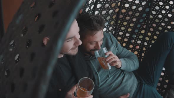 Happy Gay Couple Sitting on the Swing on the Balcony and Laughing