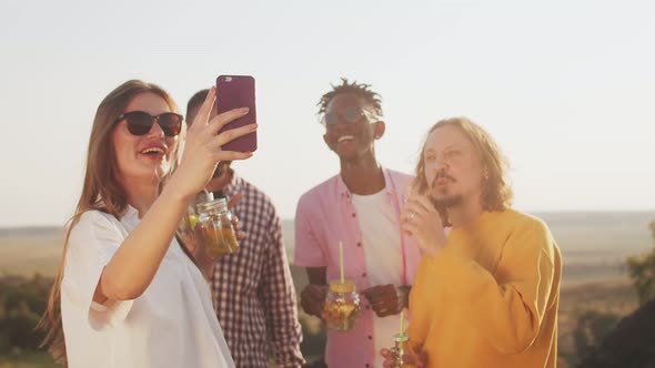 Happy emotional friends drink cocktails, laugh and woman in foreground takes selfie on her phone.