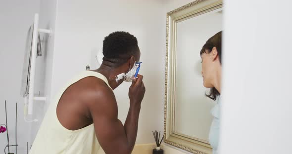 Happy diverse couple looking in mirror and shaving in bathroom