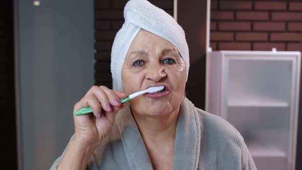 Senior Woman Grandmother with Towel Brushing Teeth and Looking Into a Mirror After Shower at Home