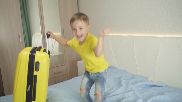 Happy Cute Boy Jumps on the Bed in Bedroom Near Packed Suitcase