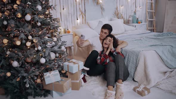 Young Couple Hugging and Taking Pictures with the Phone, While Sitting Next To the Christmas Tree at