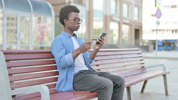 Excited Young African Man Shopping Online Via Smartphone While Sitting Outdoor on Bench