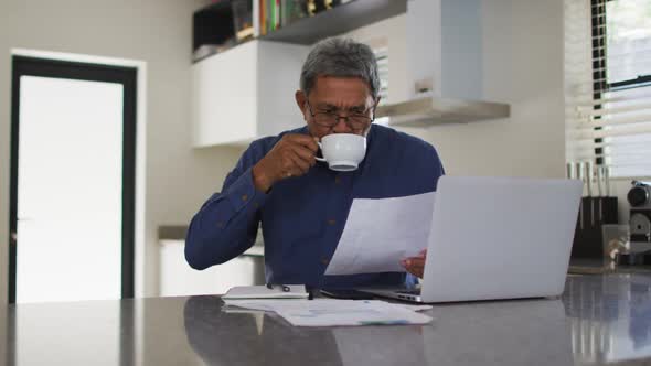 Senior mixed race man using laptop and drinking coffee in kitchen