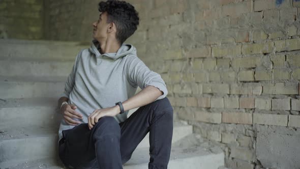 Portrait of Troubled Young Man Sitting on Stairs in Abandoned Building and Looking Around. Stressed