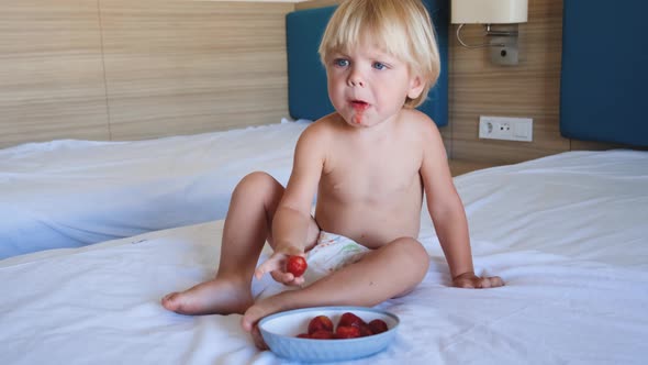 a Little Boy Sits on a Bed and Eats Strawberries