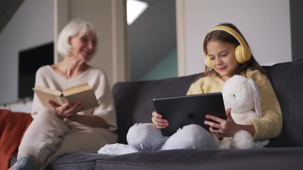 Girl looking at tablet and talking with grandmother reading book