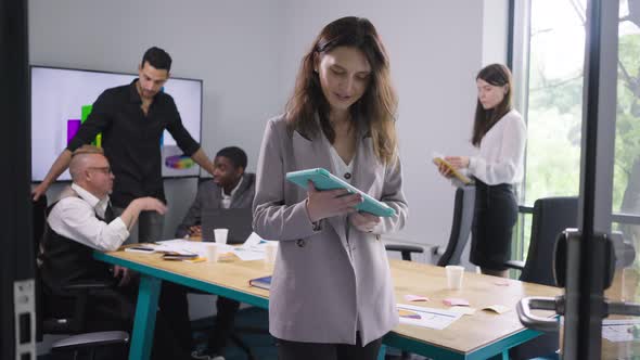 Smiling Satisfied Caucasian Woman Posing with Tablet in Office with Colleagues at Background