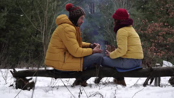 Young Man and Woman Drink Tea By the Fire in the Winter Forest