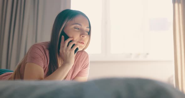 Young Attractive Happy Caucasian Woman Lying on Bed and Speaking Phone at Home in Morning