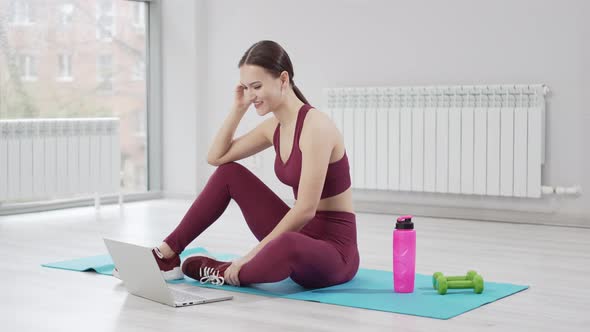 a Young Woman in a Burgundy Tracksuit with Laptop on a Mat in a Gym