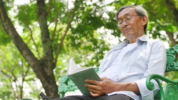 An old Asian man sits comfortably reading a book in a green park.