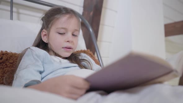 Beautiful Caucasian Girl with Grey Eyes Holding Book and Reading. Portrait of Little Lady Lying