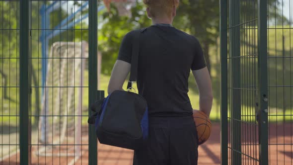 Back View of Confident Sportsman Entering Outdoor Court with Sports Bag and Ball. Young Caucasian