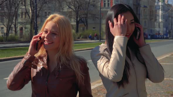 A Young Caucasian Woman and a Young Asian Woman Talk on Smartphones in a Street in an Urban Area