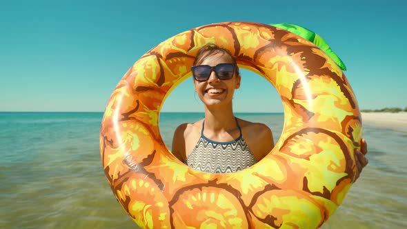 Portrait of Happy Smiling Young Woman in Sunglasses with Inflatable Pineapple Standing in Sea Water