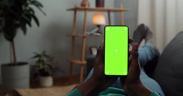 Afro American Male Person Lying on Couch and Holding Modern Phone with Both Hands, Man Browsing