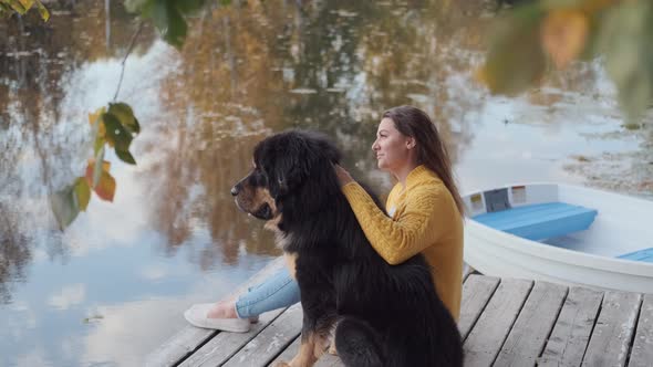A Young Woman in a Yellow Sweater Is Sitting on the Pier and Stroking a Large Dog, Next To the Boat