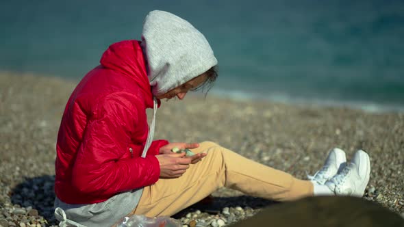 Young Woman in Red Jacket and Hoodie Sitting on Pebble Sea Beach at Windy Sunny Day and Picking Up