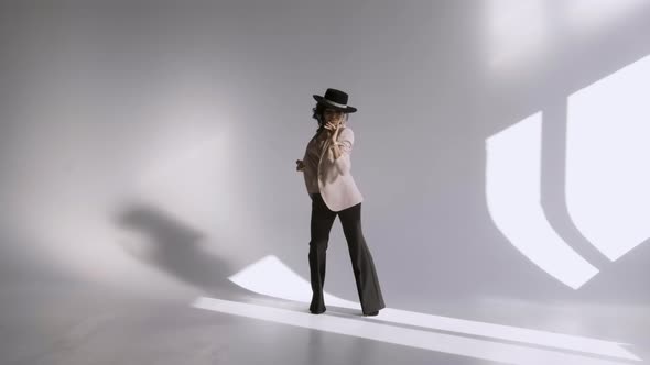 Gorgeous Young Woman in Black Trousers and a Hat Poses in a Light Studio Against the Background of a