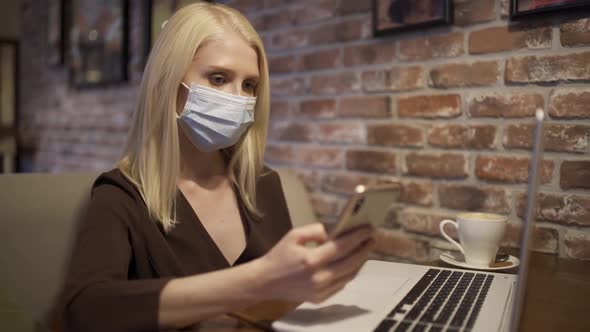 Young Working Woman in a Medical Mask Scrolls the Phone in a Cozy Cafe