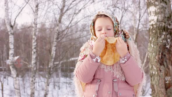 Cute girl in a traditional Russian headscarf with bagels eats pancake on winter background.