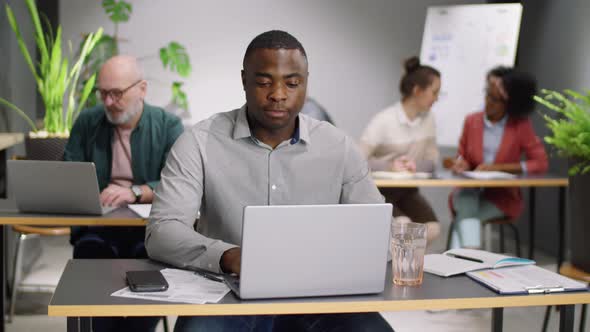 Afro-American Male Office Worker Using Laptop and Drinking Water at Desk