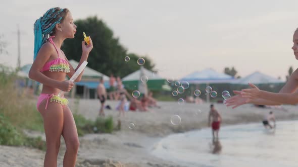 Young Girl Catches Bubbles on the Shore of the Lake Which Her Little Sister Lets Go